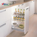 Referencing Soft-closing 3-layer pull out kitchen baskets PTJ004E5-150.jpg, a slim three-tier chrome wire pull-out basket is fully extended from a white kitchen base cabinet, neatly organizing tall seasoning bottles, stacked canned goods, and ceramic bowls on its distinct shelves.