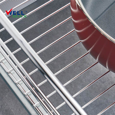 Referencing Kitchen drawer basket PTJ007V.jpg, the chrome drawer basket is pulled out from a white cabinet on sliding tracks, organizing white plates, bowls, and a stainless steel pot. Additionally, Kitchen drawer basket PTJ007V 1.jpg provides a detailed close-up of the polished chrome wire structure situated next to a red cooking pan.
